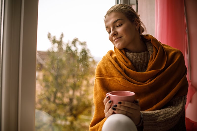 woman sitting against window, looking cozy, with a fall background.